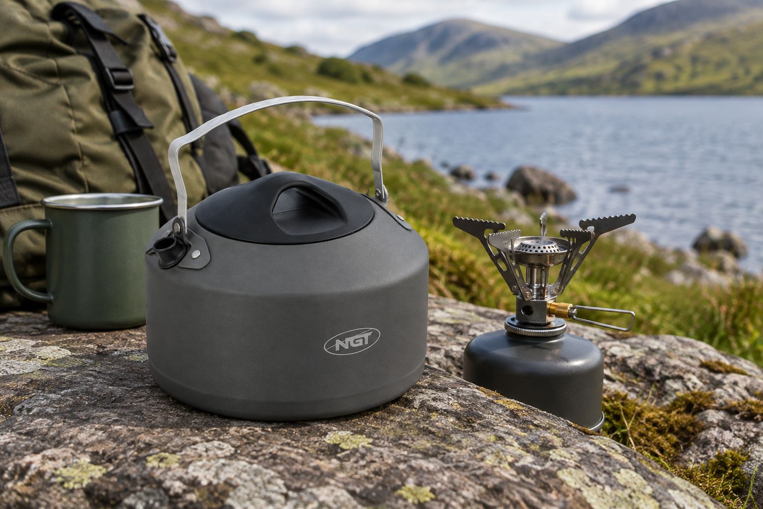 Camping kettle beside an unlit stove, mug and rucksack near a lakeshore