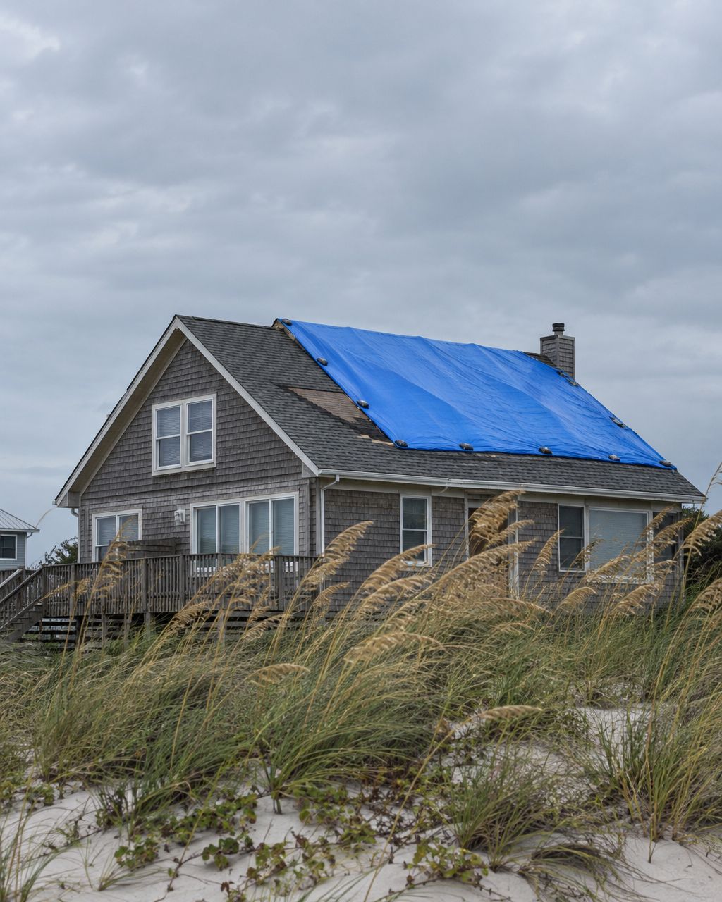 Coastal home with a blue tarp on its storm-damaged roof behind sand dunes