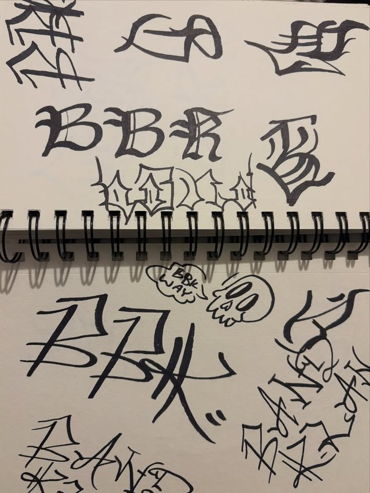 Open spiral-bound sketchbook with black hand-lettering studies and a skull doodle, with binding shadows across the paper.
