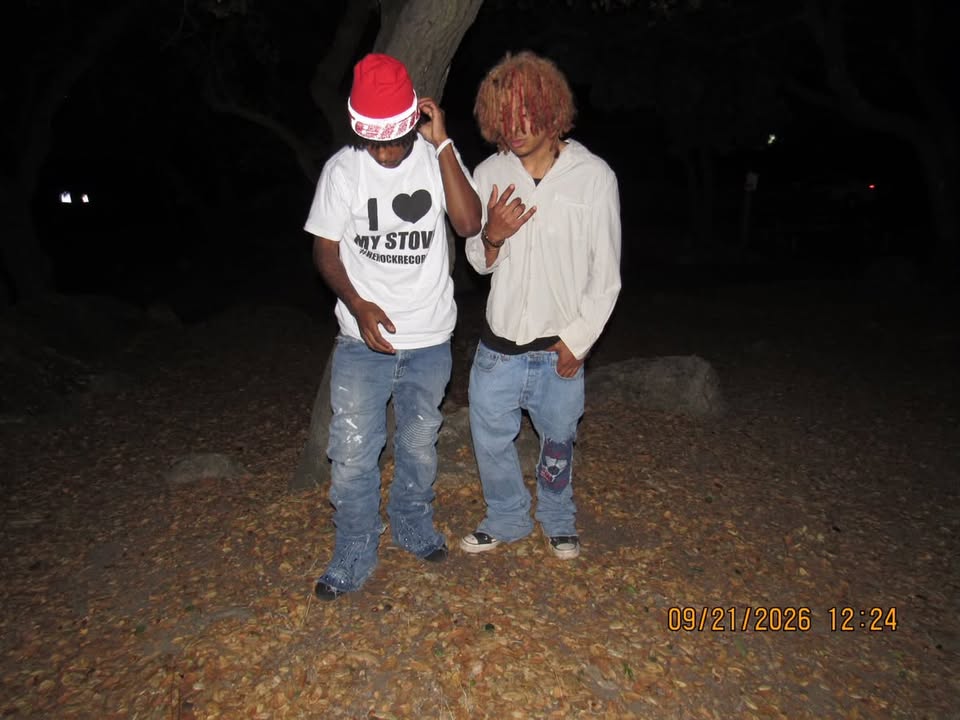 Two people standing beside a tree at night, one in a red hat and printed white T-shirt and the other in a white shirt and patched jeans.